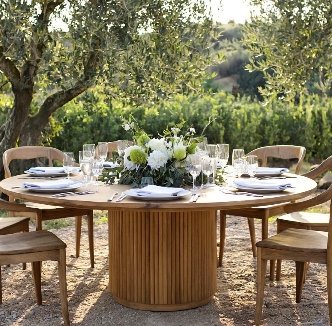 Tavolo rotondo in legno con base a doghe verticali allestito per pranzo all'aperto sotto ulivi mediterranei, con sedie coordinate, centrotavola floreale bianco e calici di vino